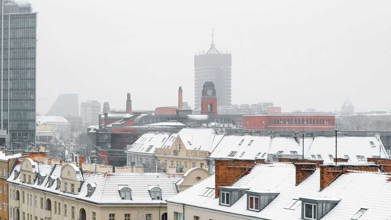 Zimowa panorama Poznania z pokrytymi śniegiem dachami i charakterystycznymi wieżowcami w tle.